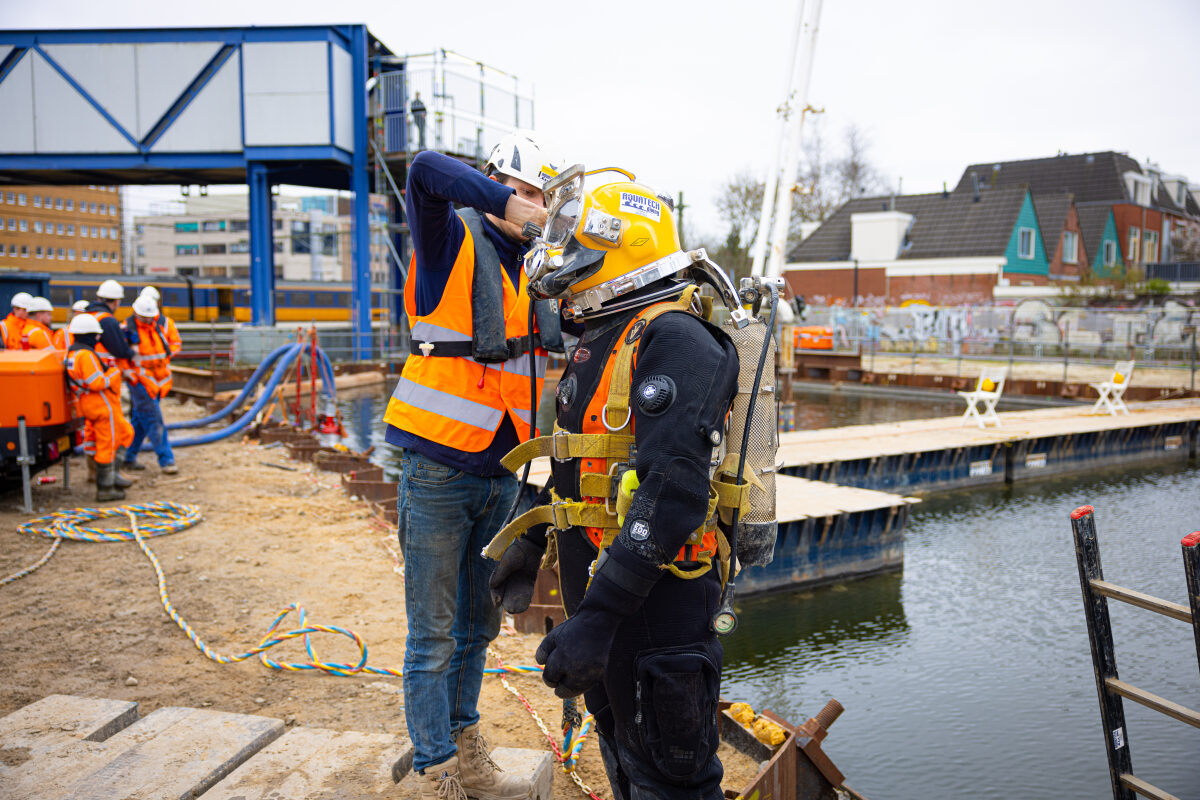 Een duiker wordt door een collega gecontroleerd voordat hij te water gaat bij het storten van onderwaterbeton voor de bouw van de bustunnel bij station Groningen